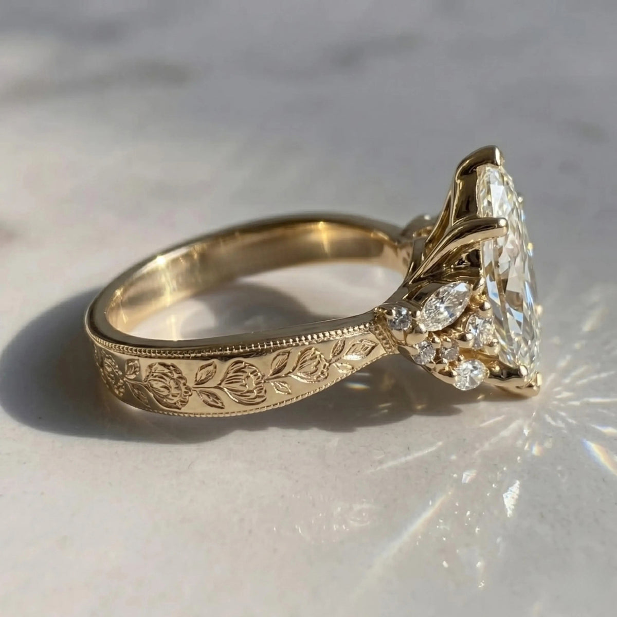 An antique-style yellow gold ring with floral engravings on the band and a large oval-cut diamond center stone, photographed on a light marble surface with bright sunlight and shadows.