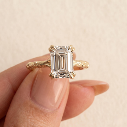A yellow gold ring with an emerald-cut clear diamond held between fingers against a soft beige background.