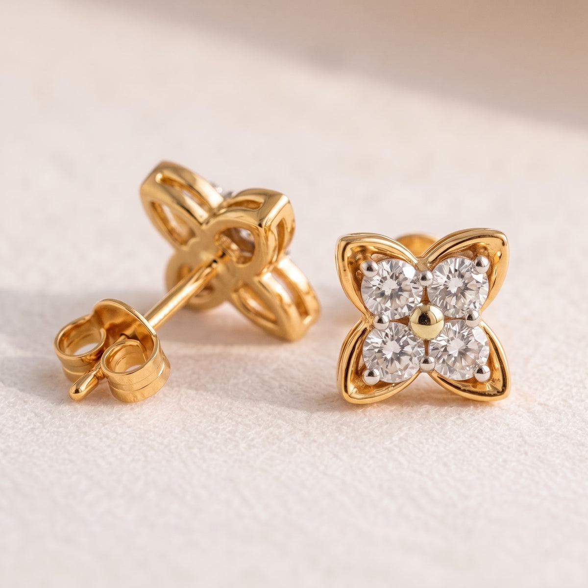 A pair of yellow gold flower-shaped stud earrings, one shown from the front with four clear round-cut stones and a central gold dot, and the other from the side/back showing the post and butterfly clasp, set against a textured off-white background.