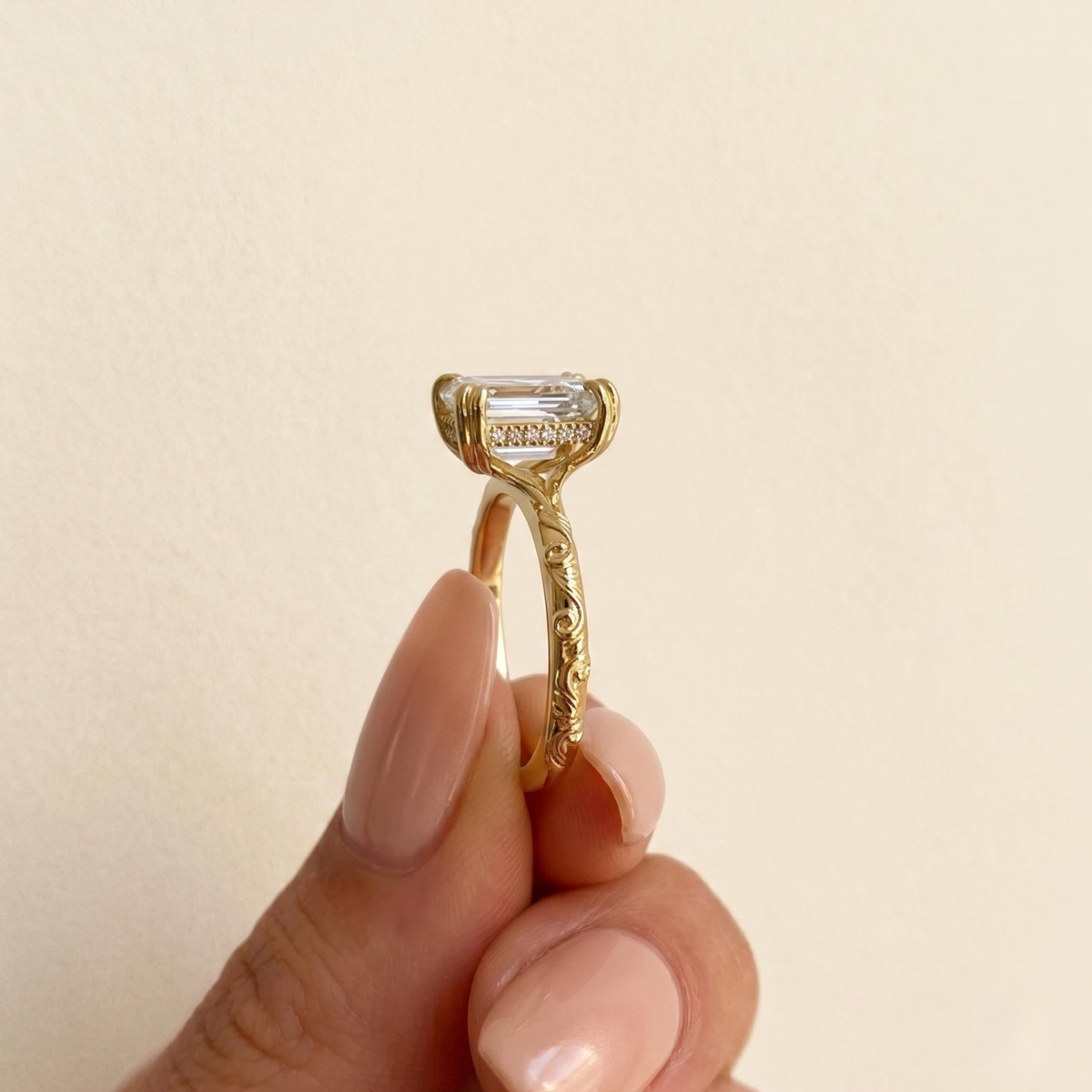 A close-up of a gold ring with an emerald-cut clear gemstone being held between fingers against a soft beige background.