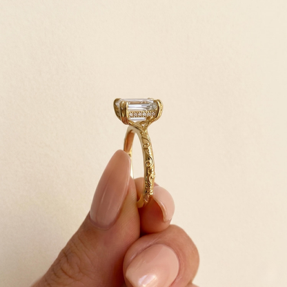 A close-up of a gold ring with an emerald-cut clear gemstone being held between fingers against a soft beige background.