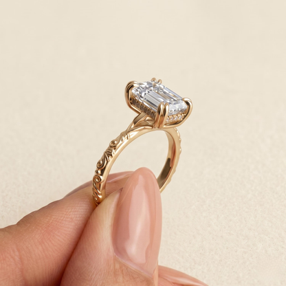 A close-up of a gold ring with an emerald-cut clear gemstone being held between fingers against a soft beige background.