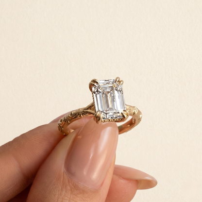 A close-up of a hand holding a gold ring with an emerald-cut clear gemstone against a light beige background.