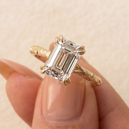 A close-up of a gold ring with an emerald-cut clear gemstone being held between fingers against a soft beige background.