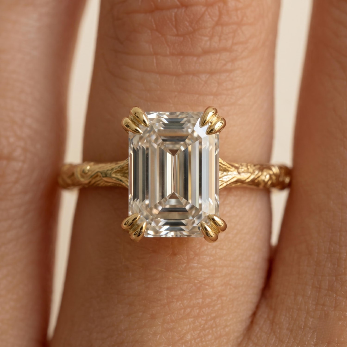 A close-up of a gold ring with an emerald-cut clear gemstone worn on a finger. The ring features a textured gold band and claw-style prongs holding the rectangular clear stone.
