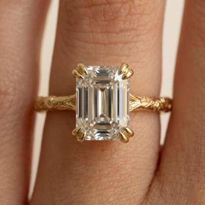 A close-up of a gold ring with an emerald-cut clear gemstone worn on a finger. The ring features a textured gold band and claw-style prongs holding the rectangular clear stone.