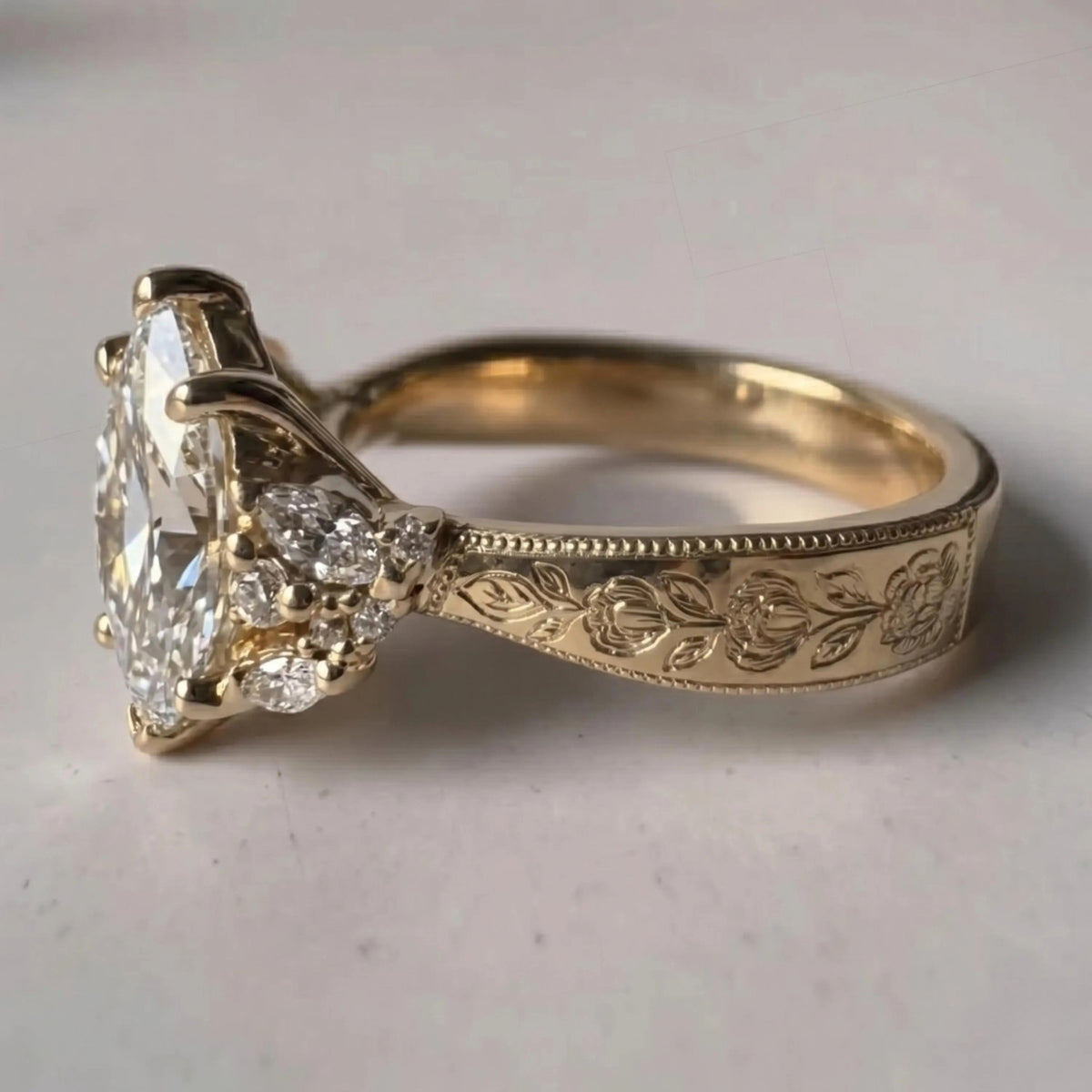 An antique-style yellow gold ring featuring a large marquise-cut diamond as the center stone, accented by small round diamonds on the shoulders and an engraved floral pattern on the band, set against a neutral light gray background.