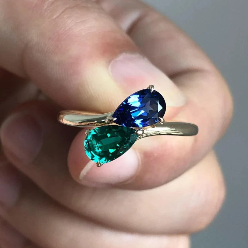 The Valencia ring showcases a stun ning 7x9mm pear-cut blue sapphire gemstone accented by pear-cut emerald gemstone, elegantly set in three modern prongs.