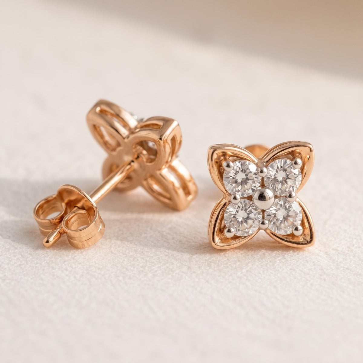 A pair of rose gold stud earrings with a four-petal flower design, one earring showing the back with the post and butterfly clutch, and the other showing the front with four clear gemstones in a diamond cluster.