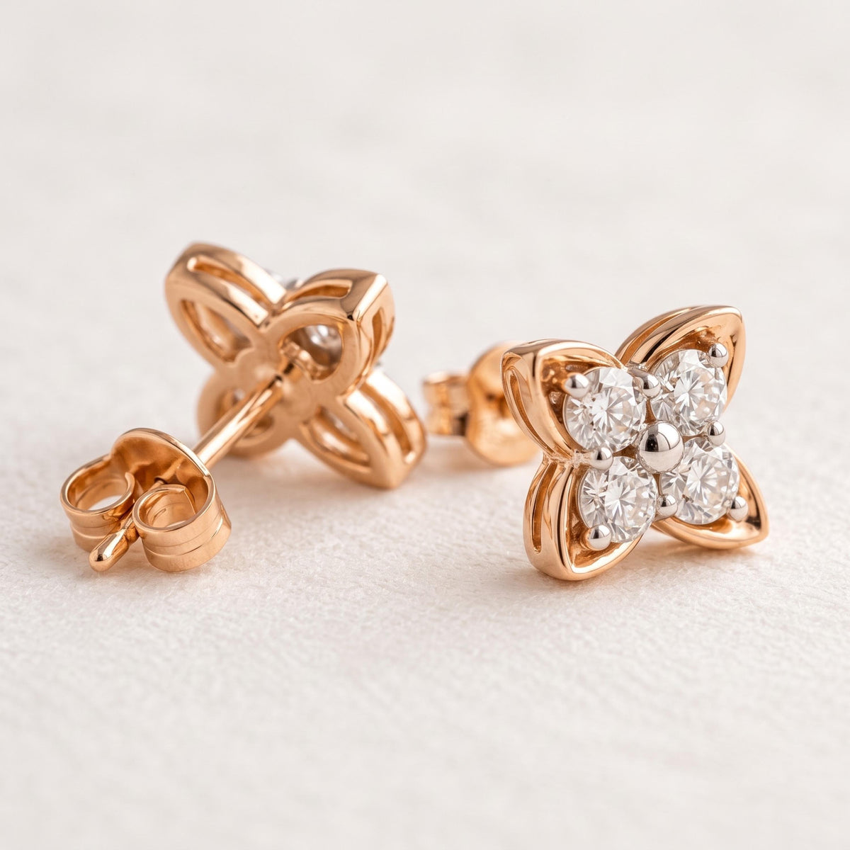 A pair of rose gold flower-shaped stud earrings, with one earring showing the post and backing and the other showing the front face adorned with four clear round-cut gemstones, set against a soft, light beige textured background.