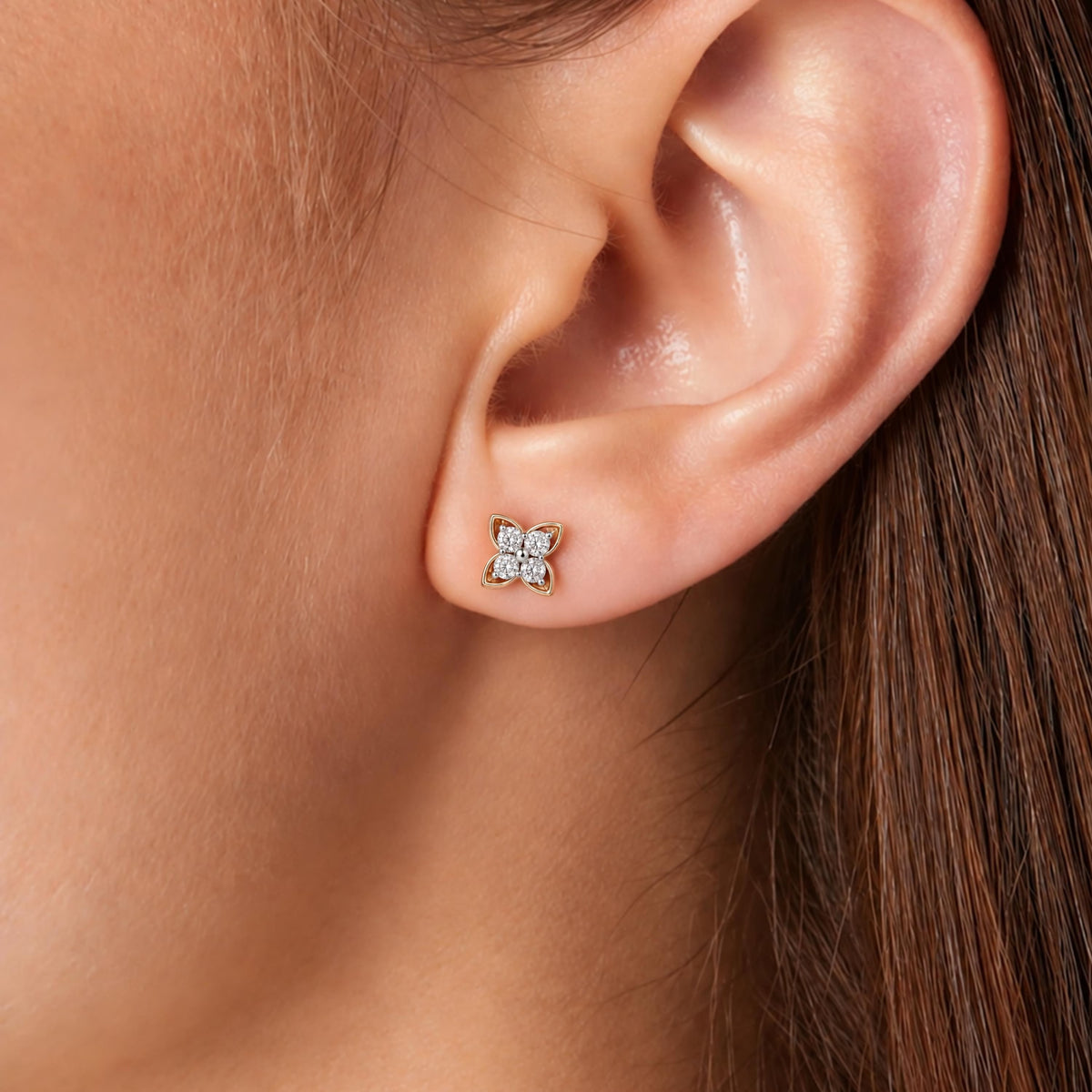 A close-up of a person's ear wearing a small gold stud earring with four clear gemstones arranged in a square.
